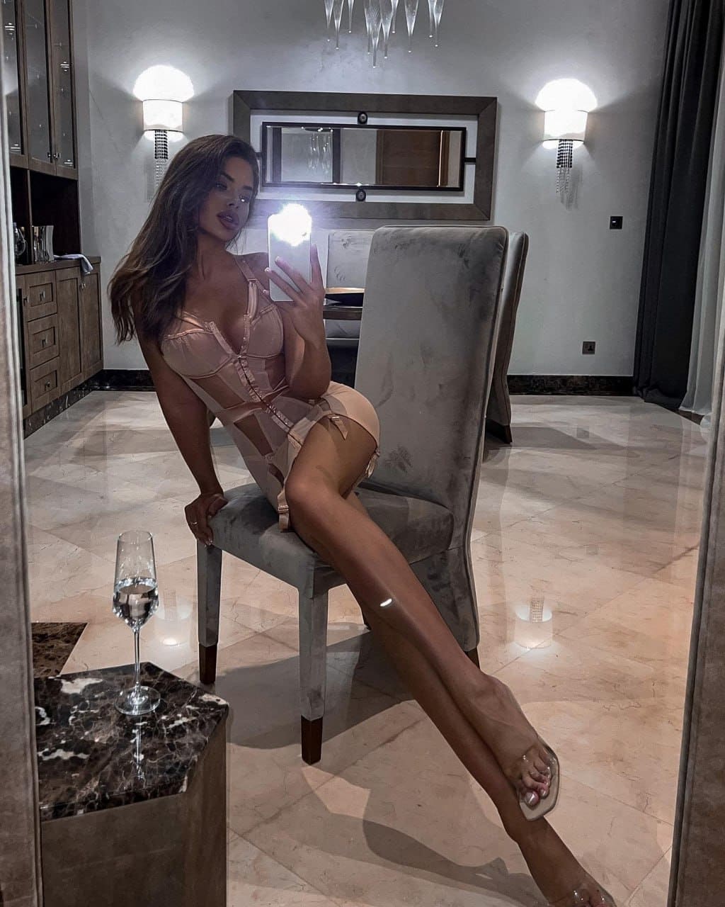 Woman seated on a high-backed chair in a marble room, wearing a nude cutout bodysuit and holding a phone with flash.