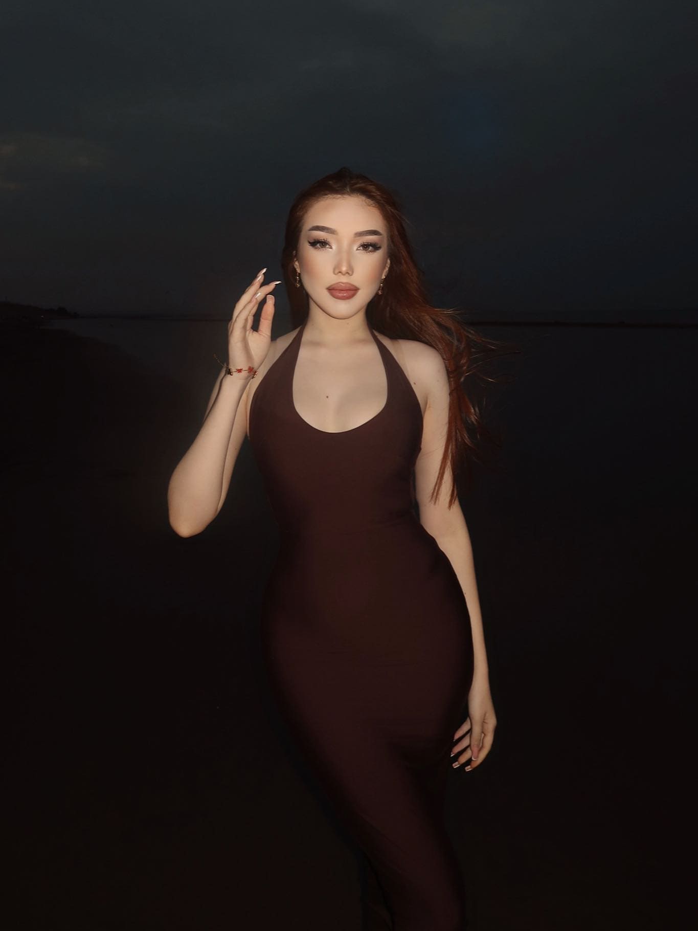 A woman stands outdoors at dusk in a dark brown halter dress, raising a hand near her face against a dark sky and water background.