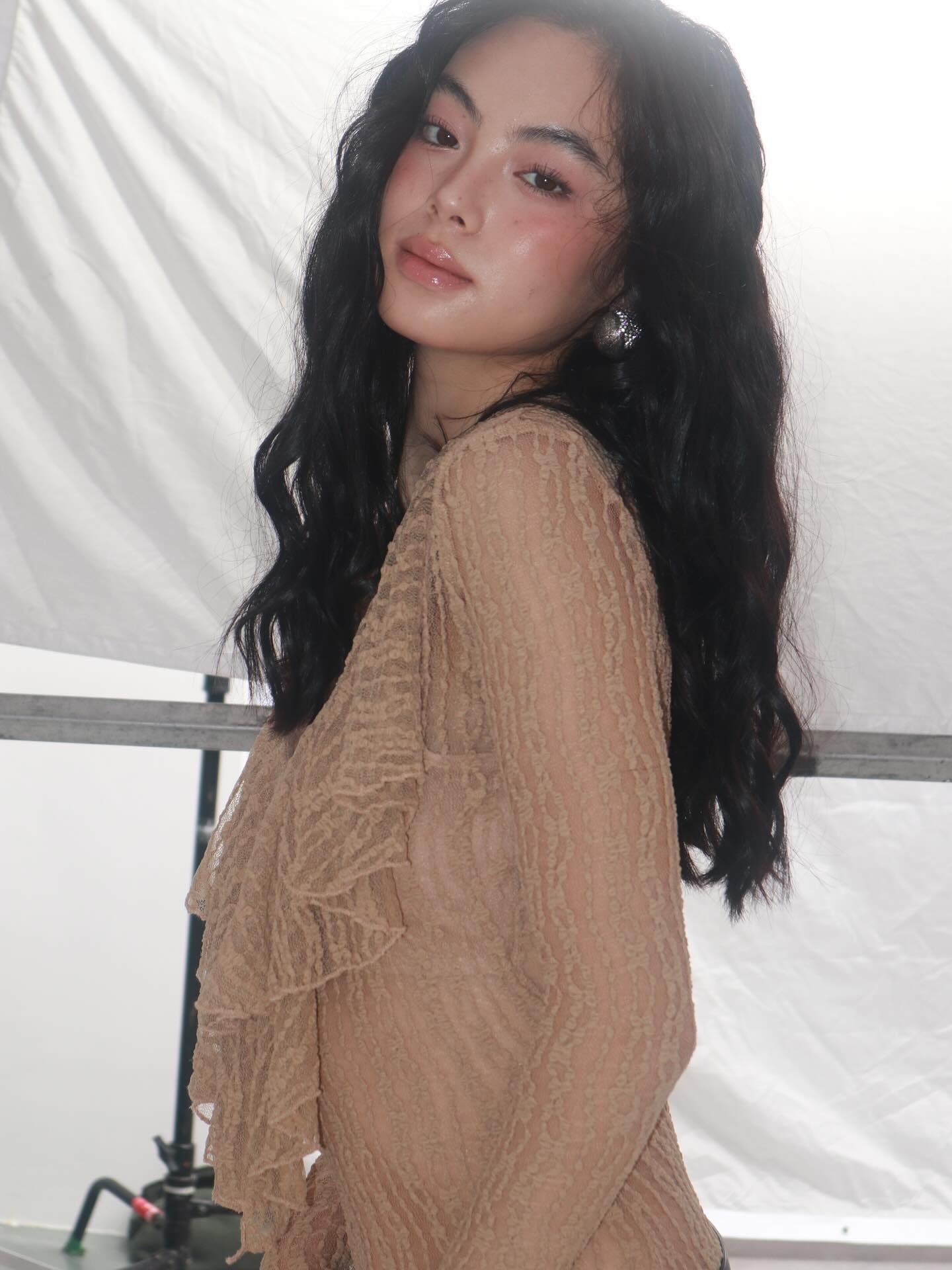 Facing left in a studio, she wears a sheer beige lace top with ruffled sleeves and a large earring.