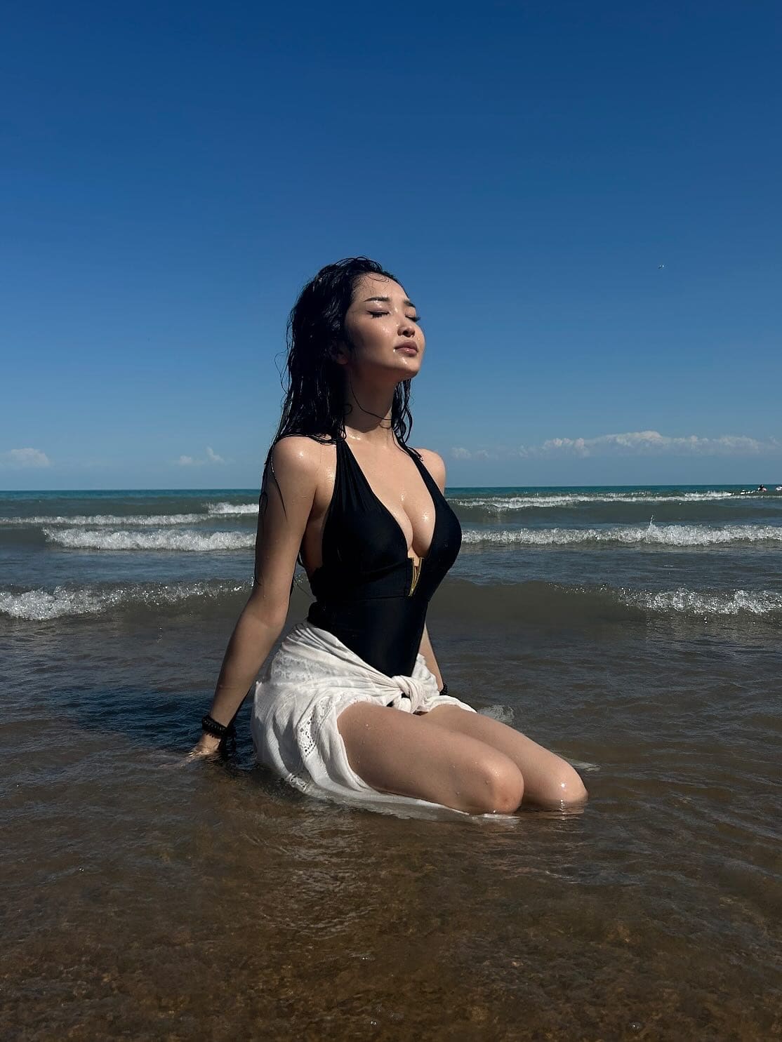 A woman with wet hair kneels in shallow ocean water, wearing a black swimsuit and white wrap skirt.