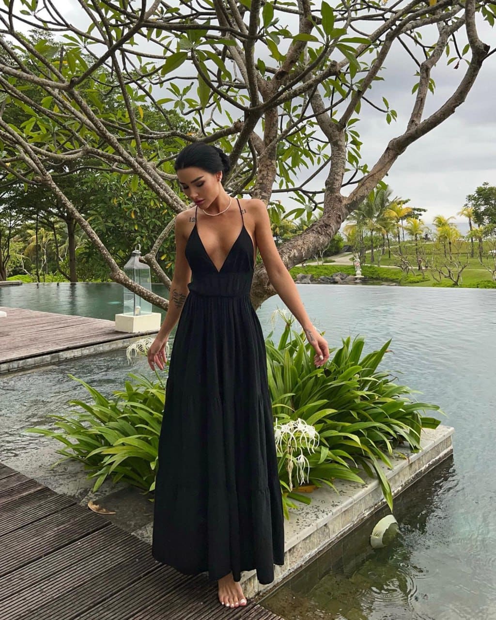 A woman stands barefoot on a wooden deck by a pool in a long black sleeveless dress, surrounded by tropical greenery.