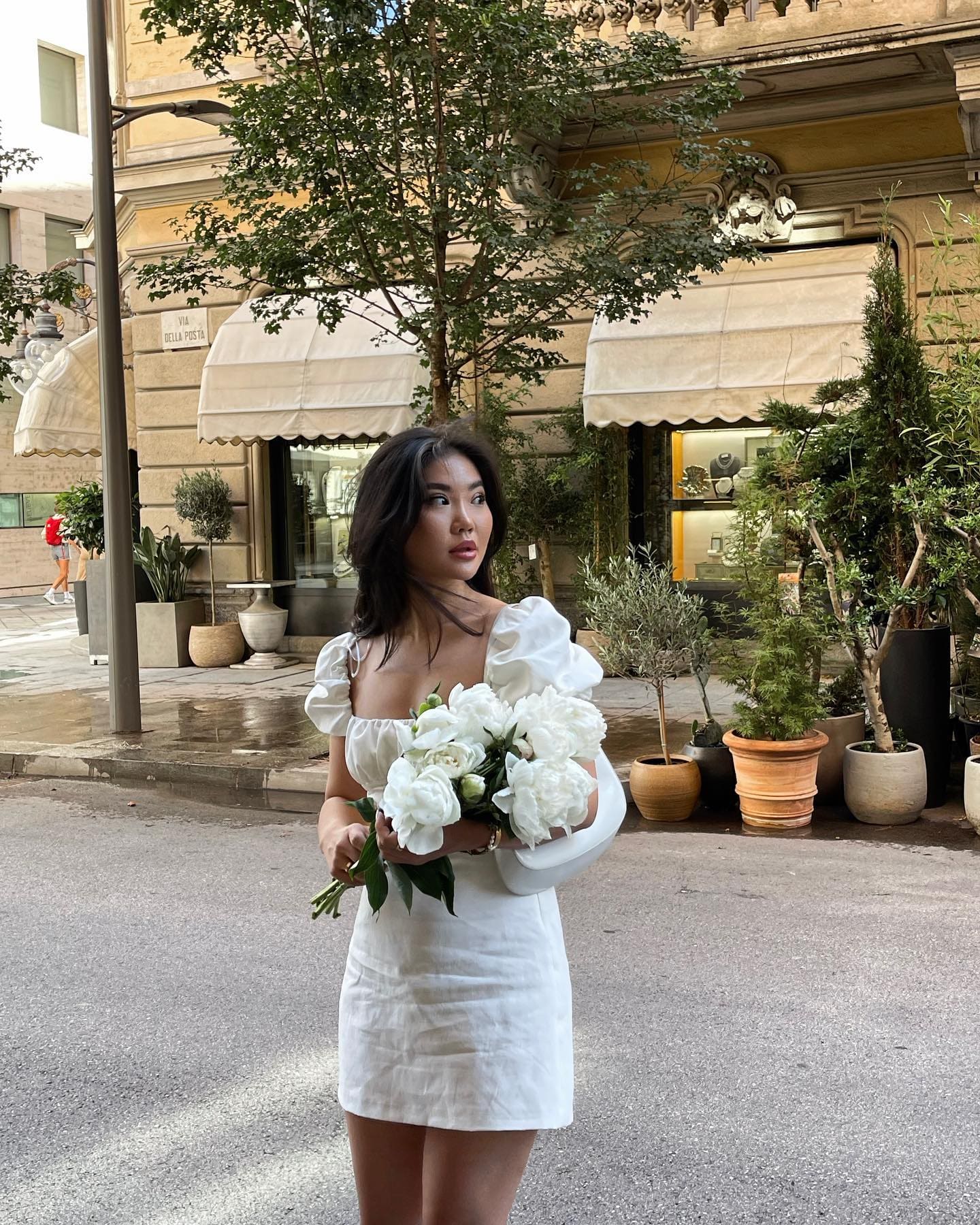 Standing on a city street holding white flowers, wearing a short white dress with puffed sleeves near awnings.