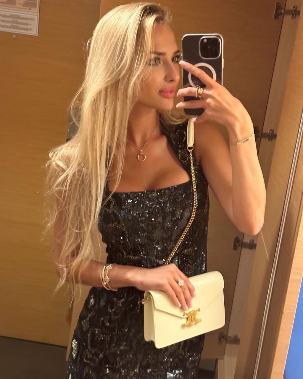 Mirror selfie of a woman in a sequined black dress with a low neckline, holding a cream chain-strap handbag in front of a wooden door.
