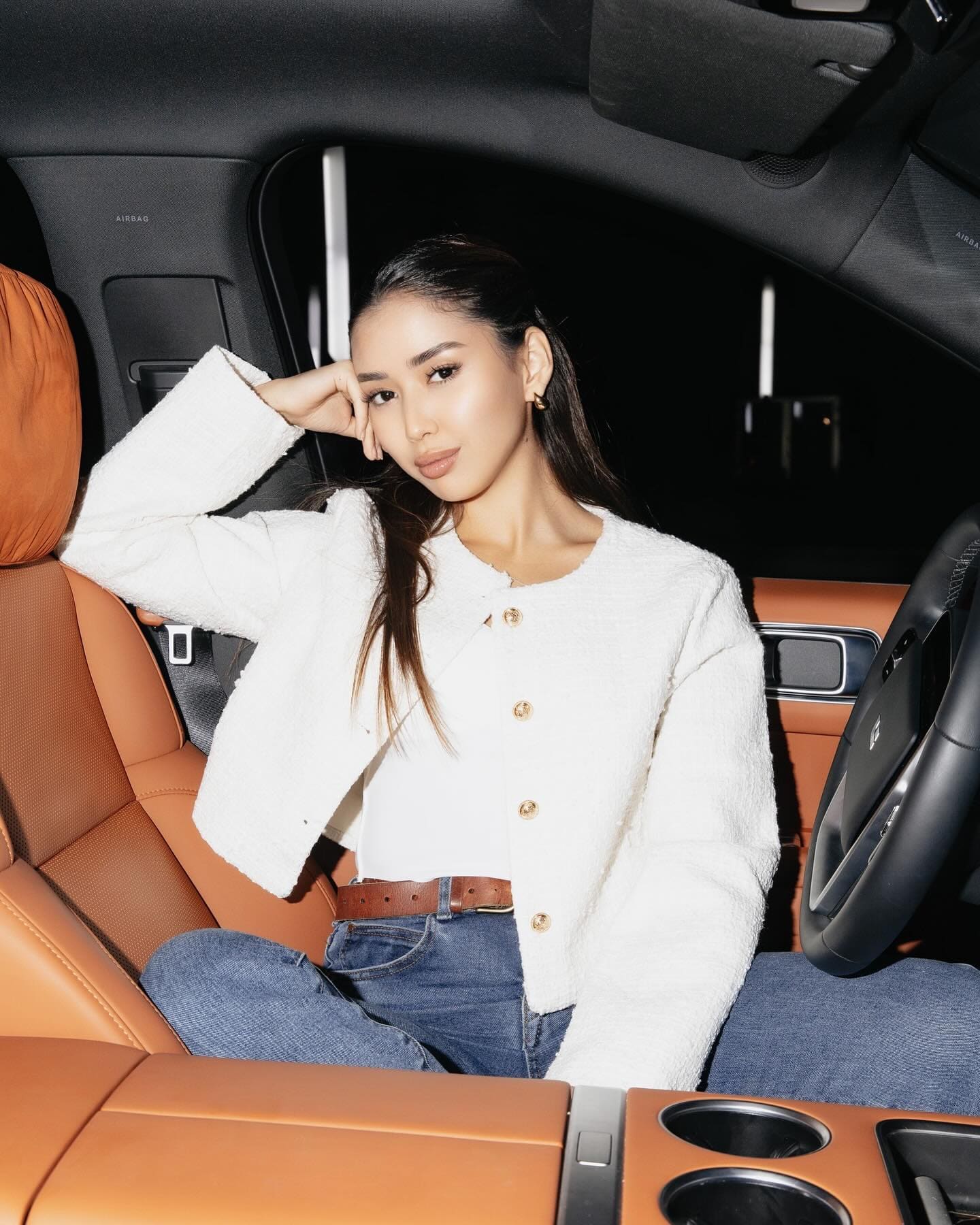 A woman sits in a car driver's seat wearing a white textured cardigan, blue jeans, and a brown belt, with a hand to her head.
