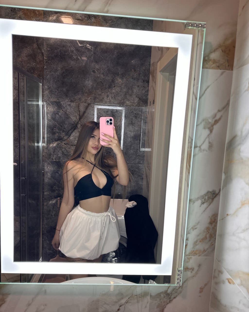 A mirror selfie shows a woman in a black bralette and white skirt holding a pink phone in a marble bathroom.