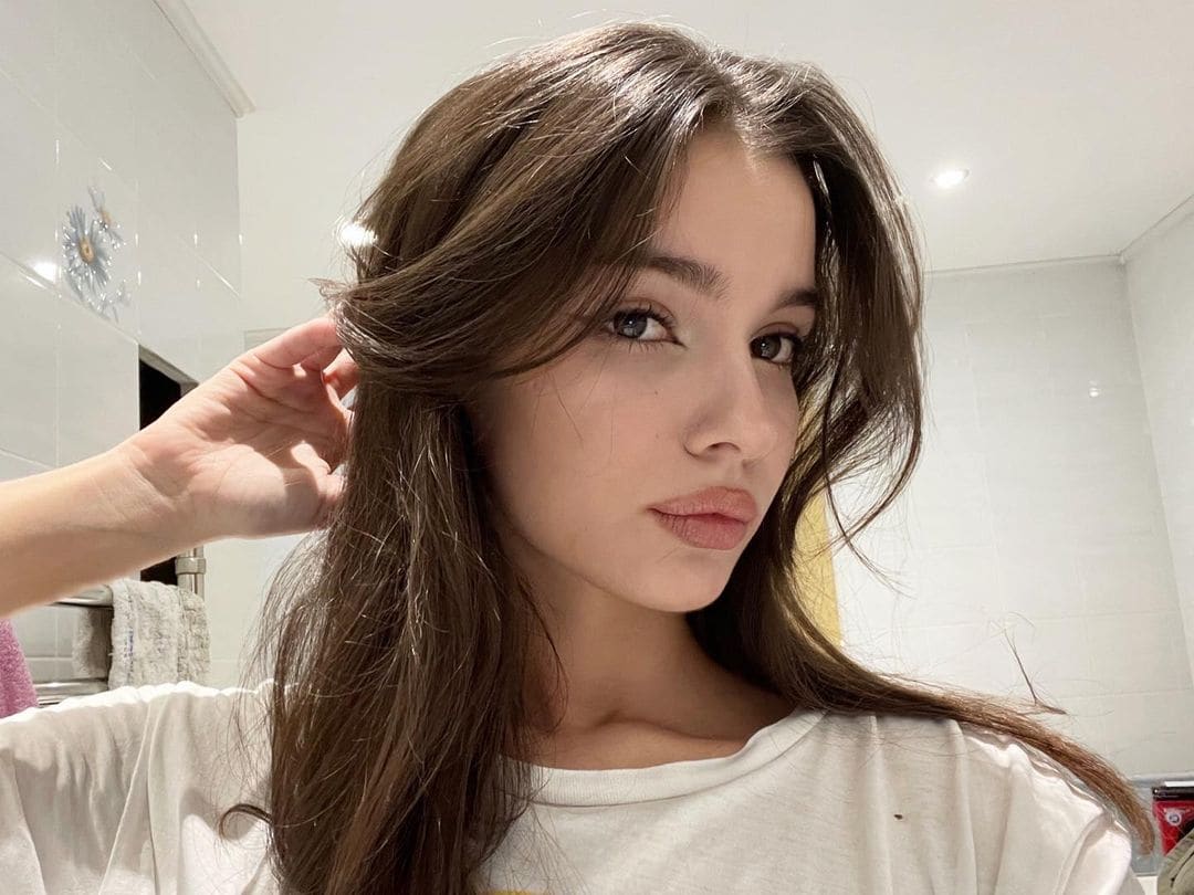Zoe takes a mirror selfie in a tiled bathroom, wearing a white t-shirt and holding a strand of her long brown hair near her ear.