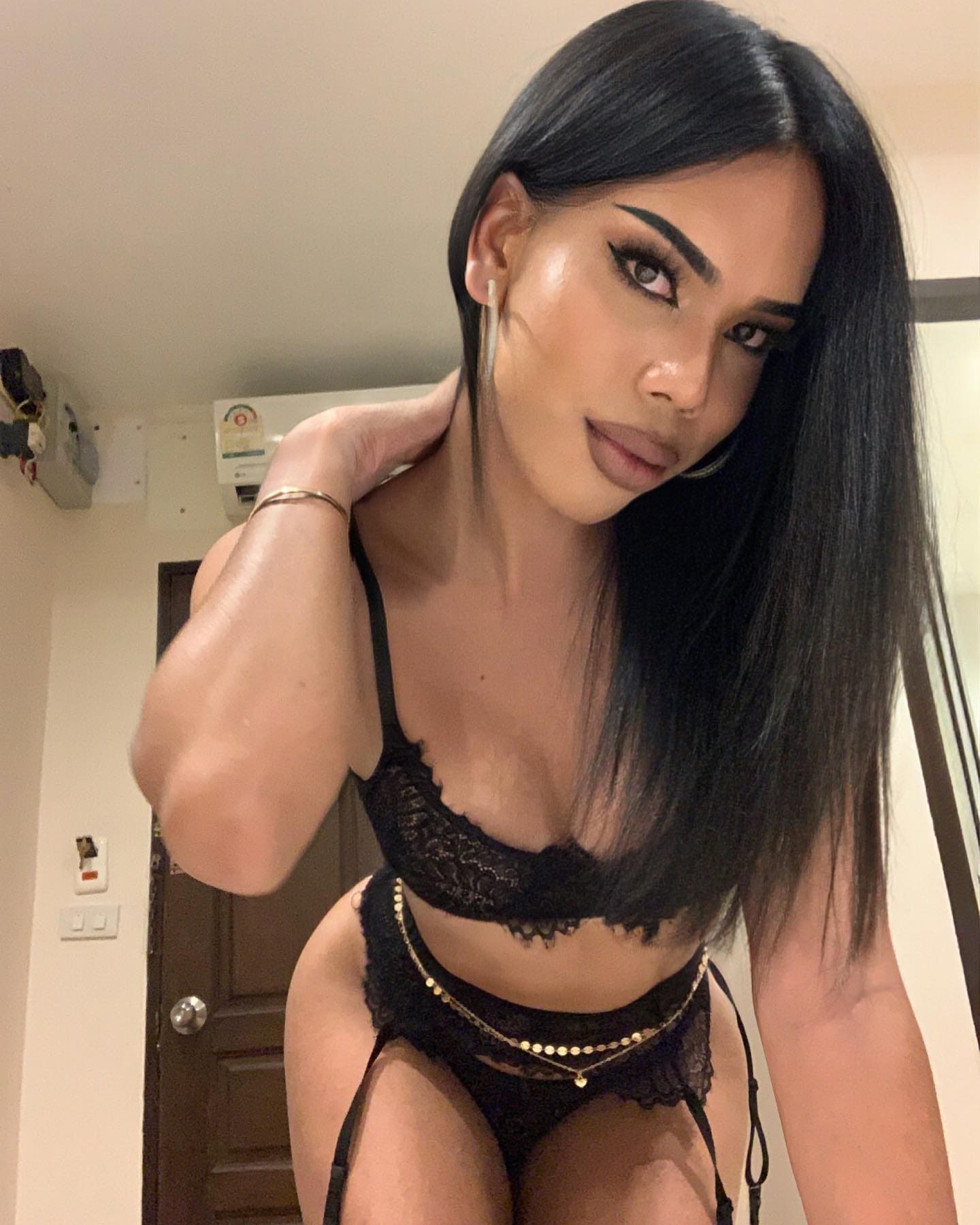 A woman in black lace lingerie and a gold chain belt rests a hand on her neck in a room with visible wall fixtures.