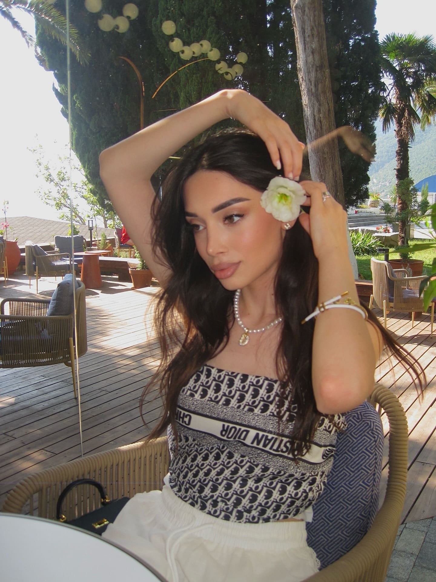 A woman in a strapless top and white pants sits in a wicker chair on a deck, holding a white flower near her ear.