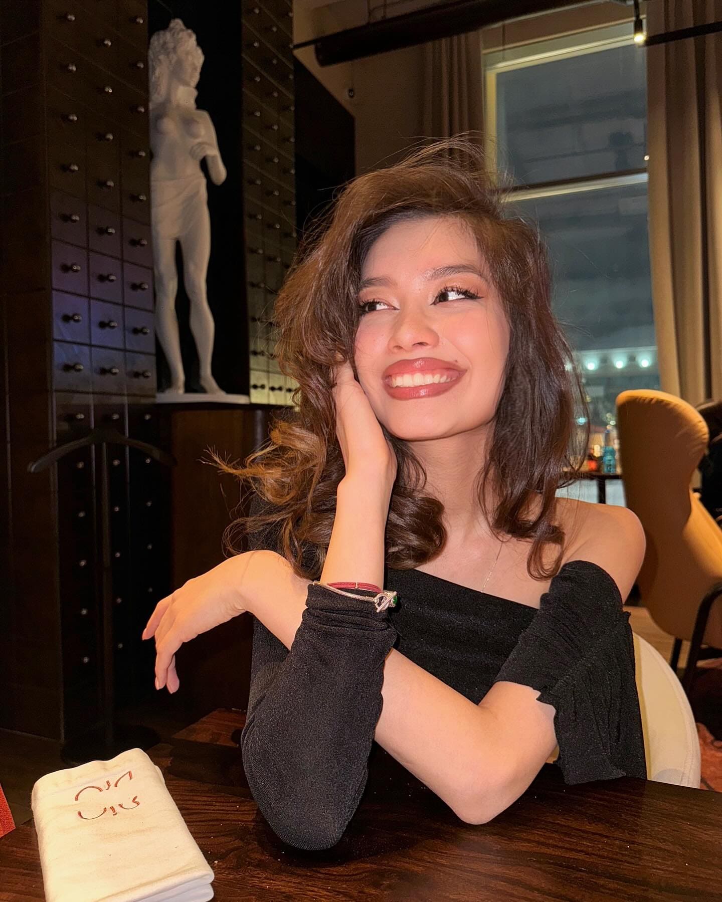 A woman sits at a wooden table indoors, wearing a black off-the-shoulder top, resting her hand on her cheek near a white statue.
