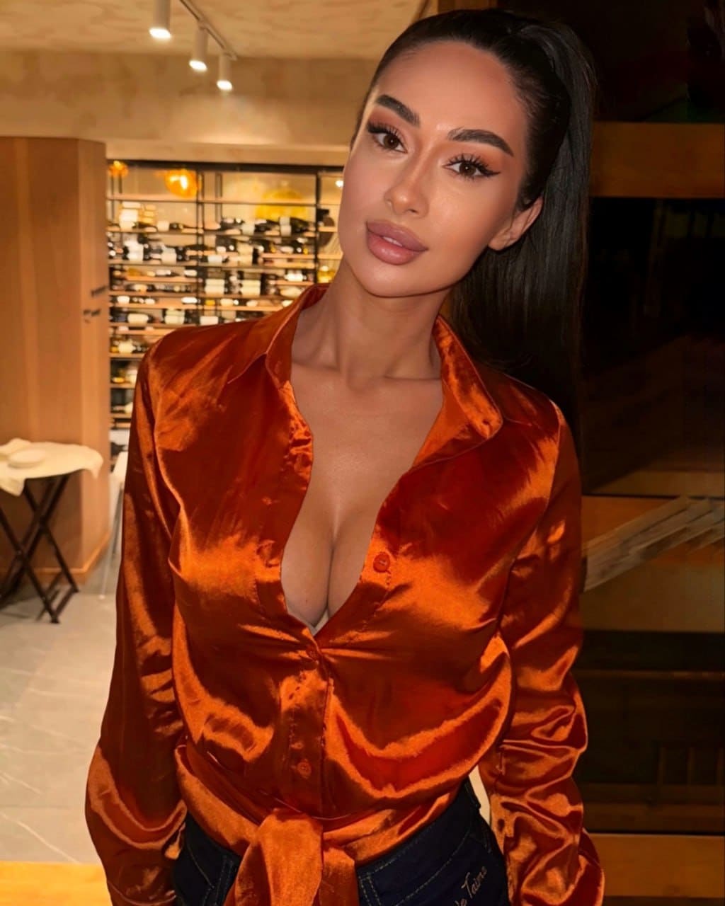 A woman in an open rust satin shirt and dark jeans stands indoors near shelves of bottles.
