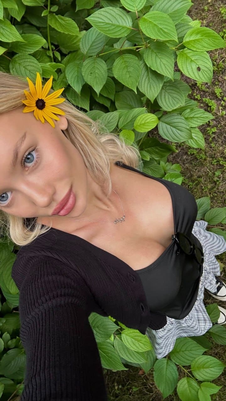 An outdoor selfie shows a woman in a black cardigan and striped skirt, with a yellow flower behind her ear and plants behind.