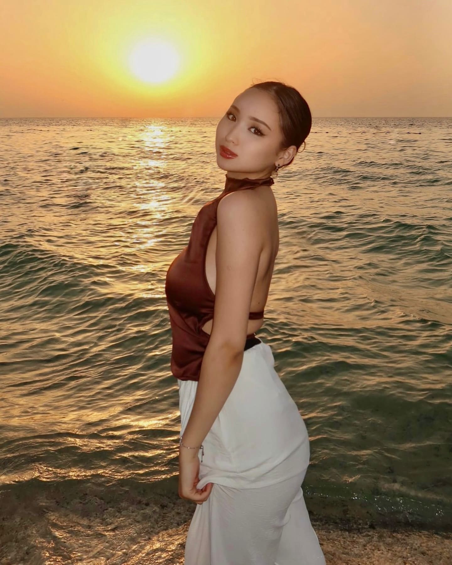A woman in a brown halter top and long white skirt stands in profile on a beach at sunset, with the sun low over the water.