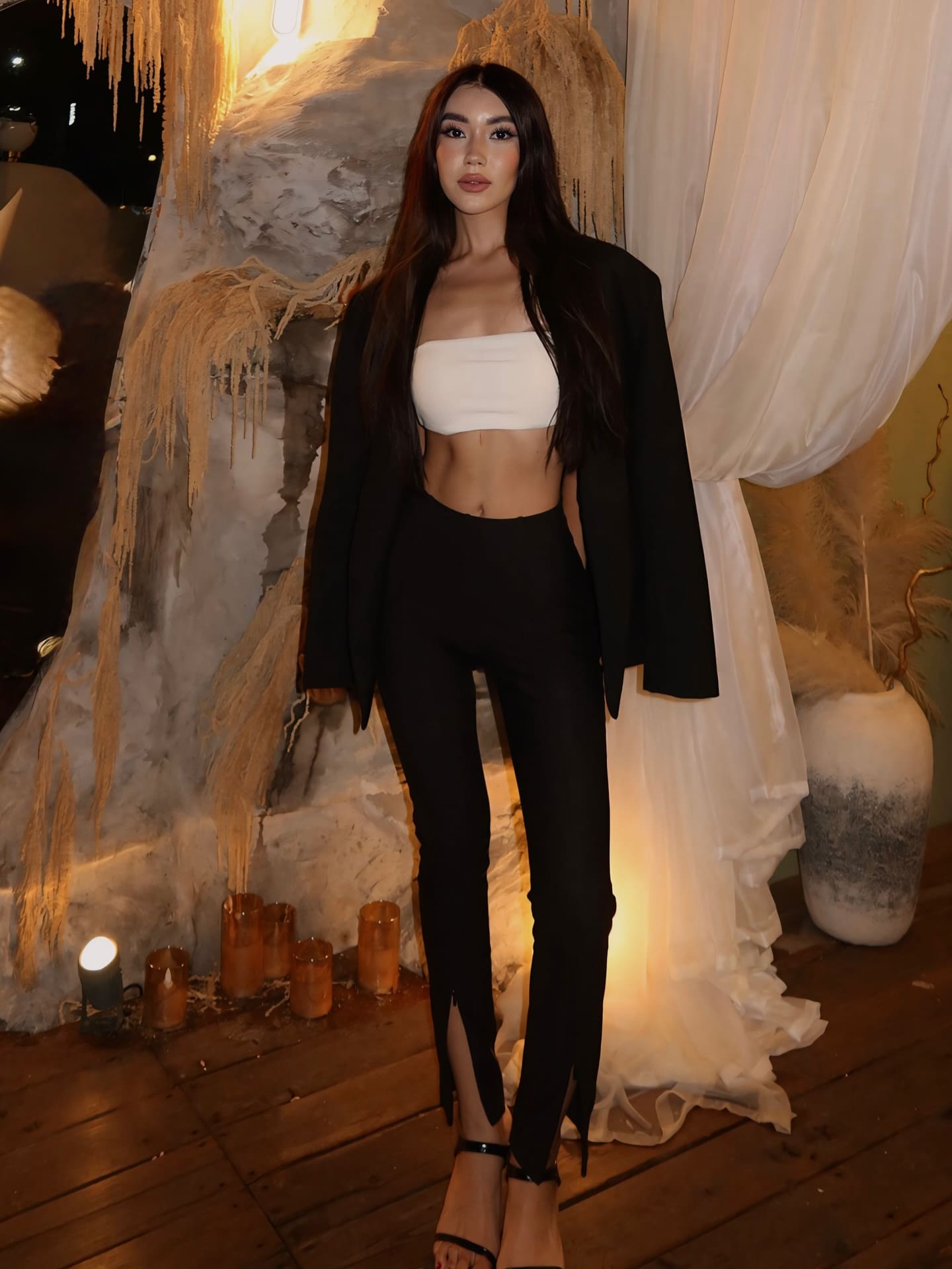 A woman in a white crop top, black pants, and open blazer stands indoors near candles and dried grass decorations in dim light.