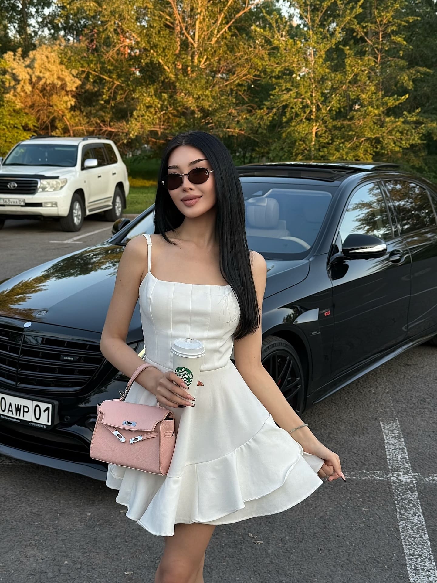 In a parking lot, she stands in a white sleeveless dress and sunglasses, holding a coffee cup and pink handbag near a black car.