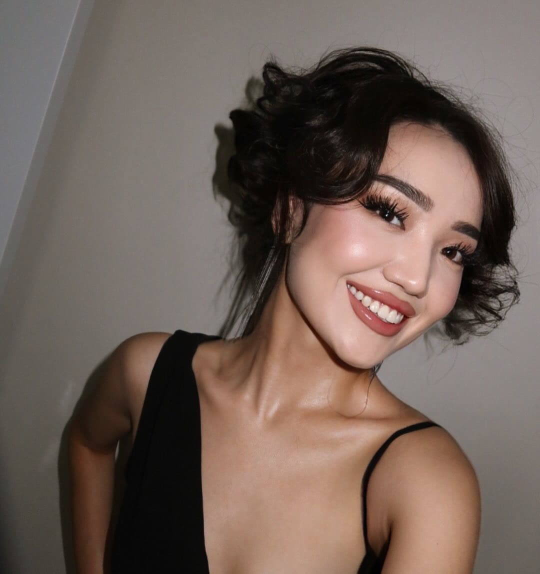 Sora smiles at the camera in a black one-shoulder top, posing against a plain light-colored wall.