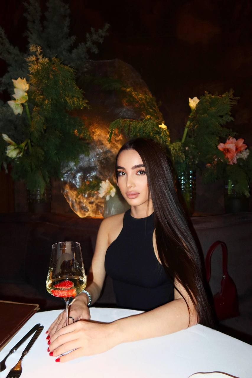 Seated at a white-clothed table, a woman in a black halter top holds a glass of white wine with a strawberry garnish in a dim room.