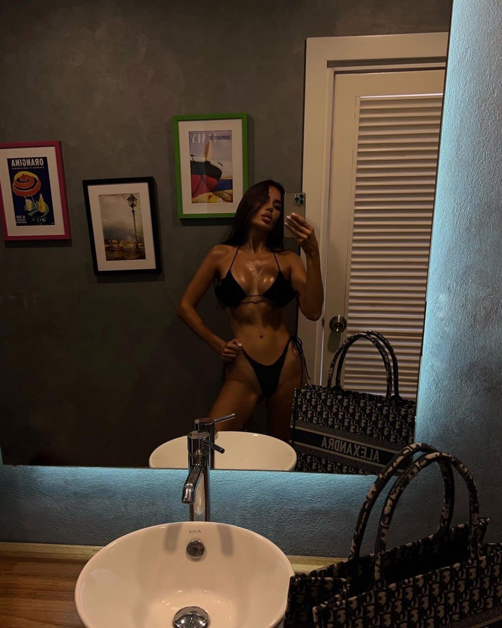 A woman in a black two-piece bikini takes a mirror selfie in a bathroom, with a white sink and patterned handbag in the foreground.
