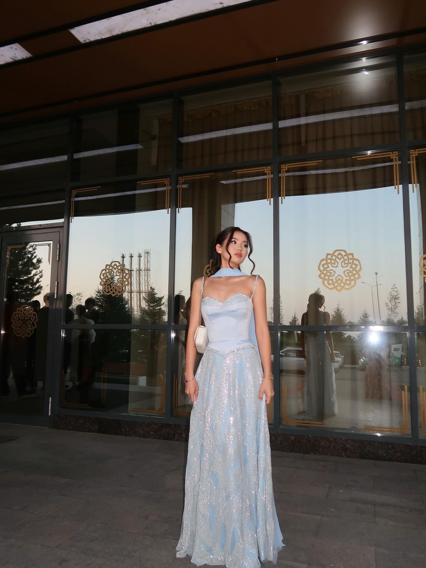 A woman in a light blue evening gown with a sparkly skirt and halter top stands before a glass facade, holding a white handbag.