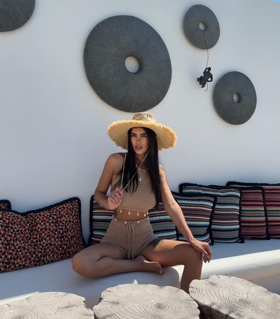 A woman sits cross-legged on a white bench wearing a tan crochet halter top, matching shorts, and a wide-brimmed straw hat, with cushions behind.