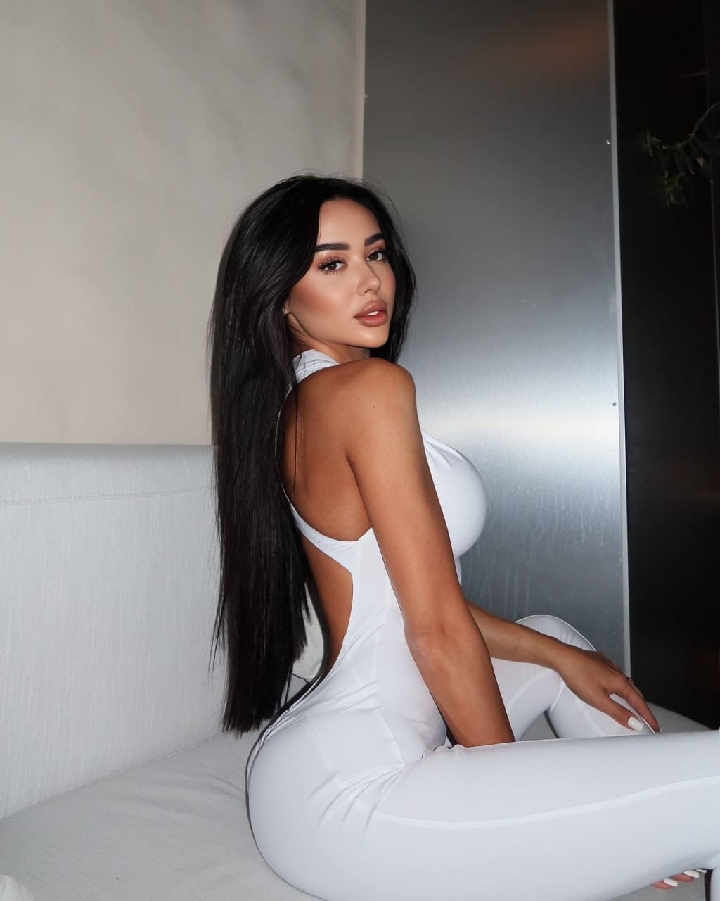 Woman in a white outfit with an open back sits on a couch, looking over her shoulder against a metallic wall panel.