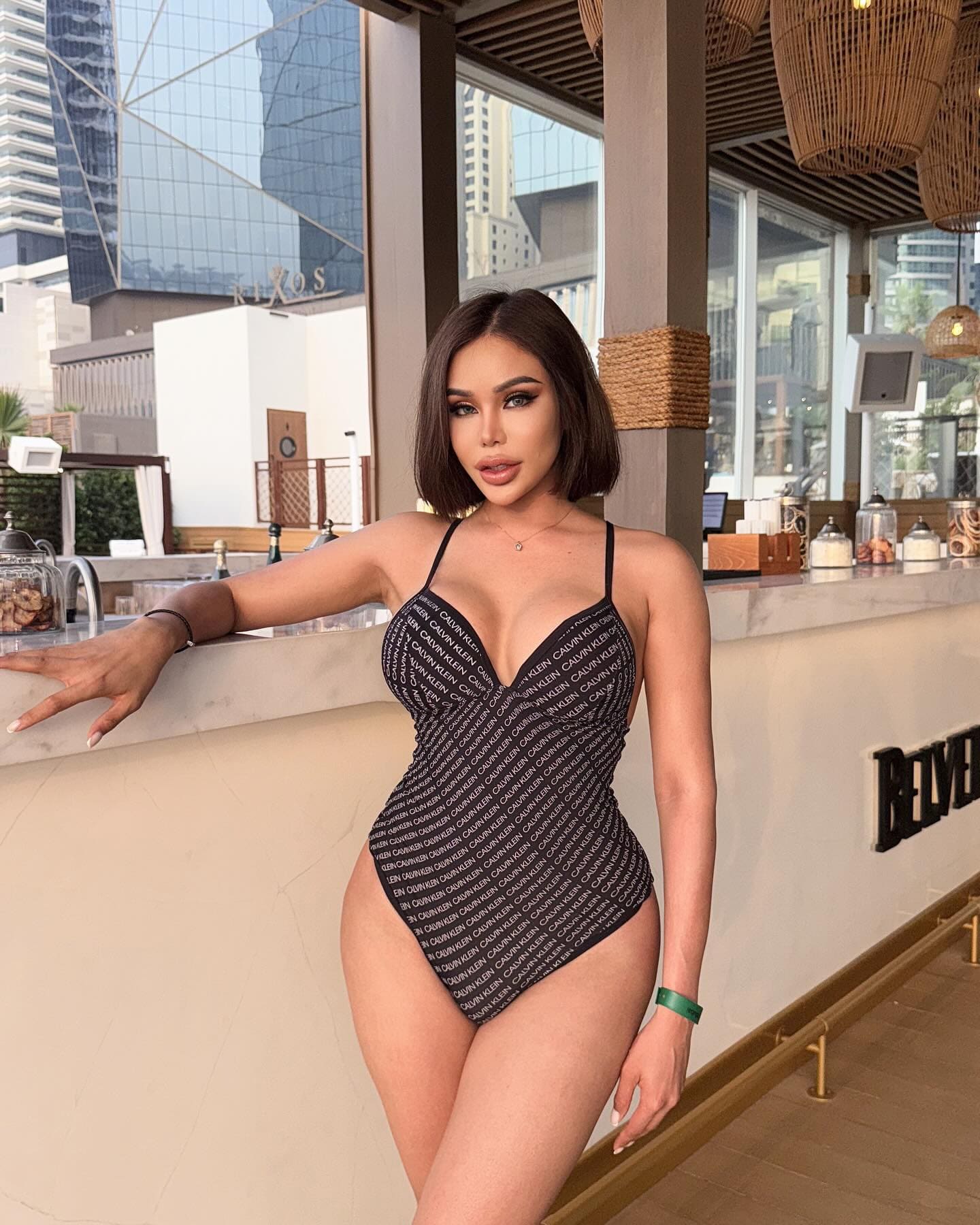 Nana stands at a bar counter in a black swimsuit with white text, resting her arm on the marble while a city skyline shows through the windows.