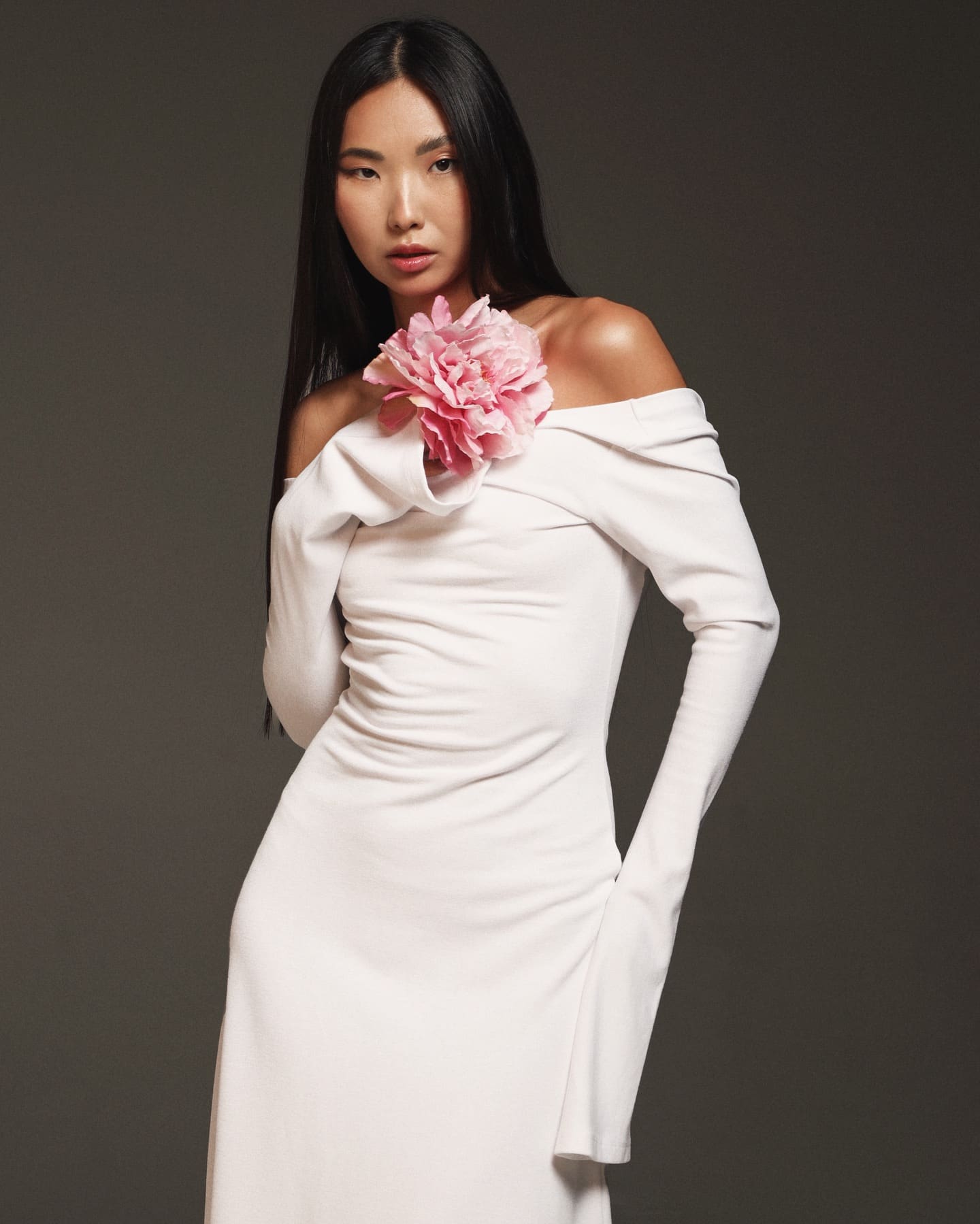 Dana stands against a gray backdrop in a white off-the-shoulder dress with a pink flower, one hand behind her back.