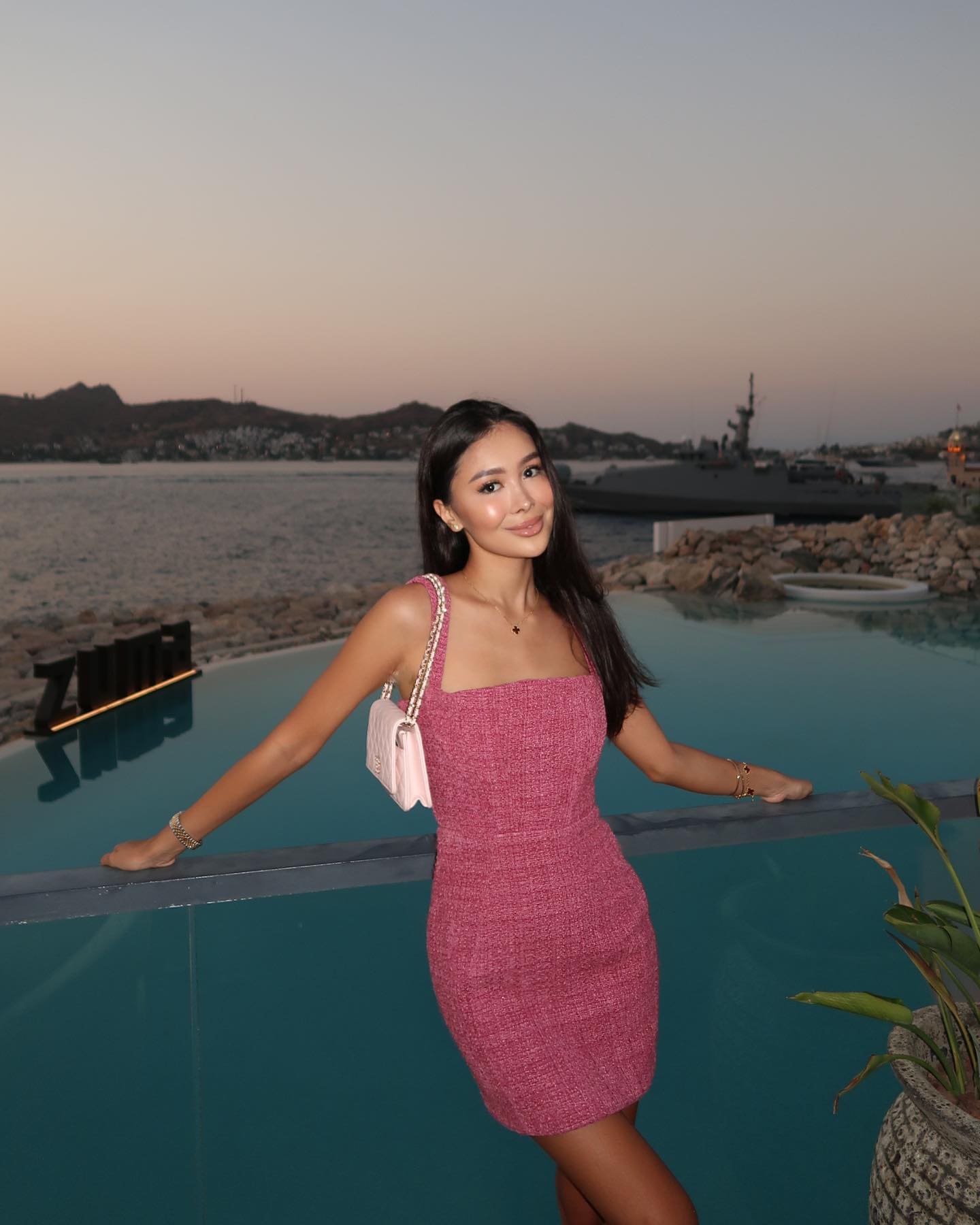 Inessa stands by an infinity pool at dusk in a pink textured mini dress and white shoulder bag, resting arms on the edge.
