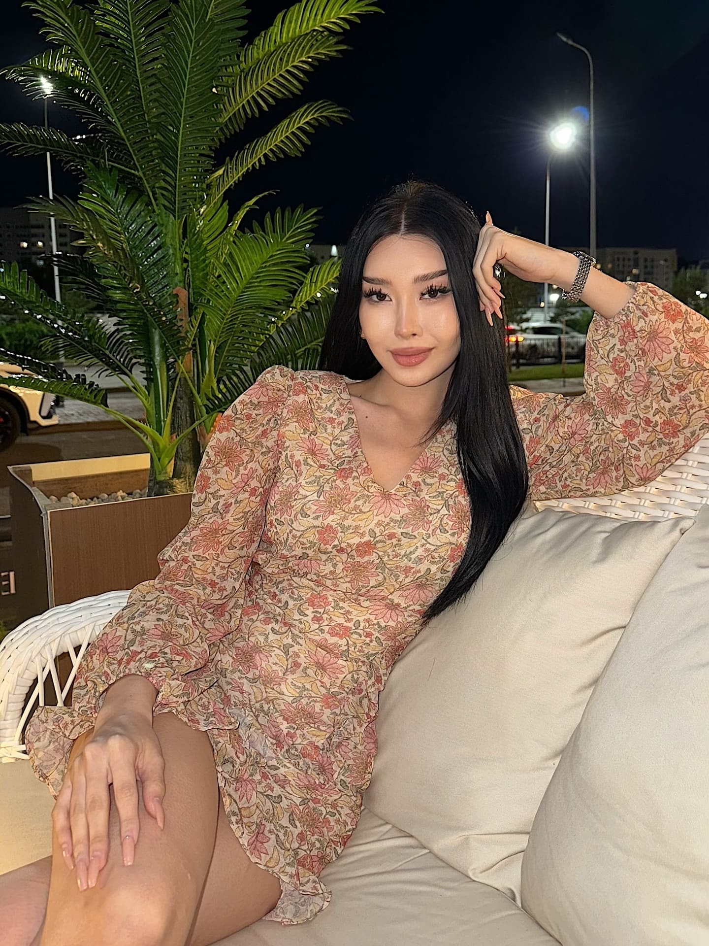 At night, she sits on a white wicker sofa in a floral dress with puffed sleeves, one hand on her head, palm plant in background.