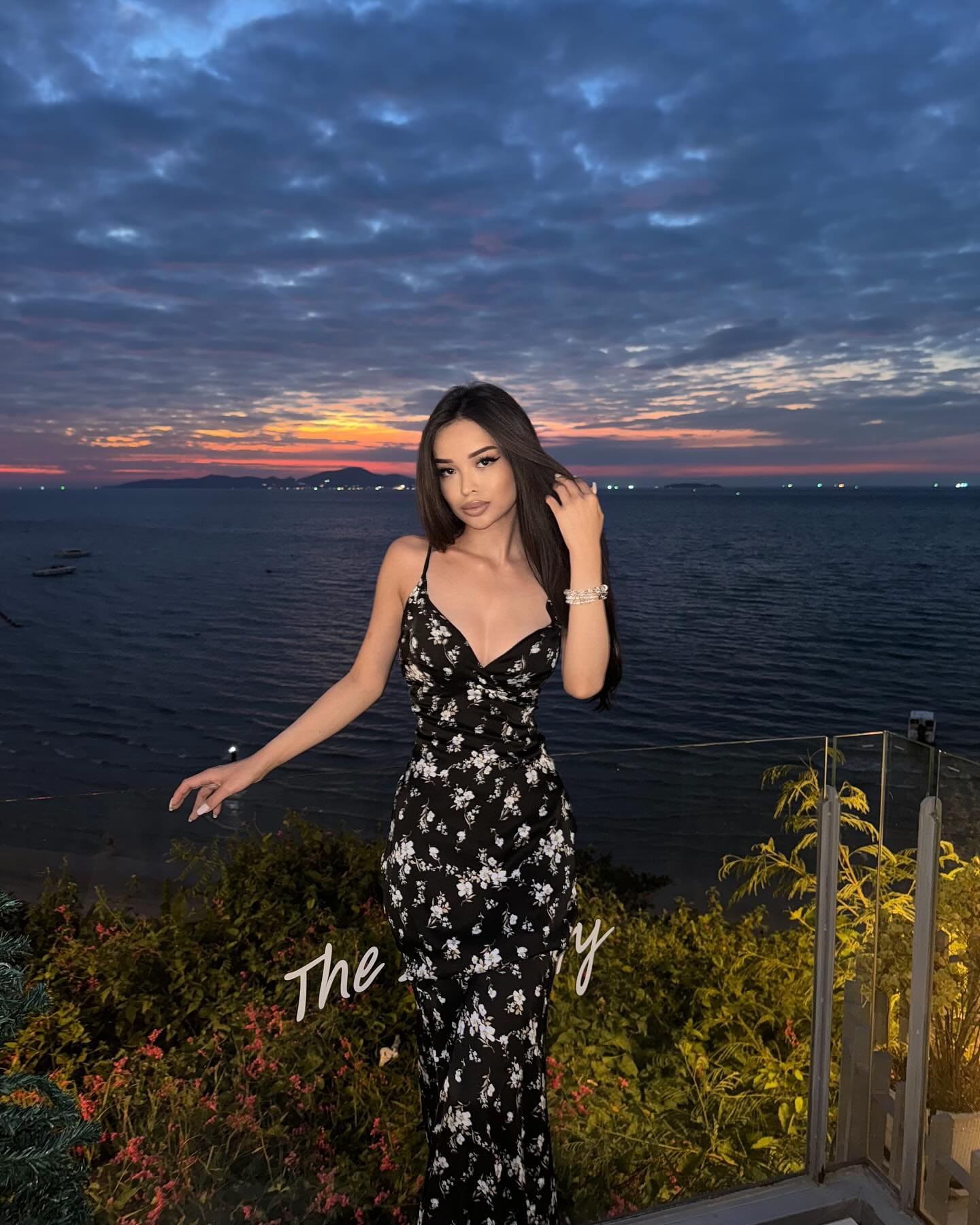 A woman in a long black floral dress stands on a balcony at sunset, one hand in her hair, overlooking the sea.
