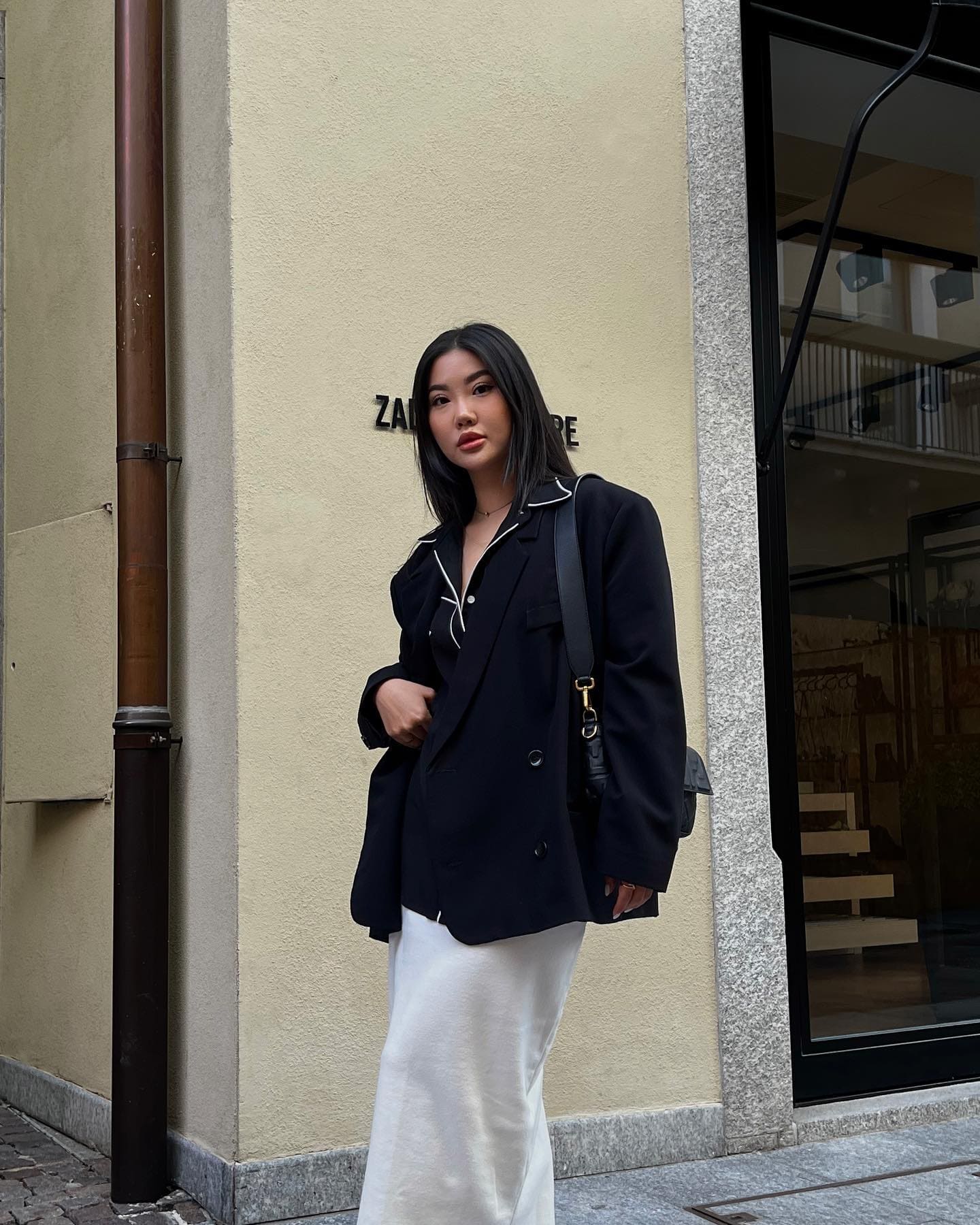 On a sidewalk before a beige building, wearing a black blazer, white top, long white skirt, and shoulder bag.