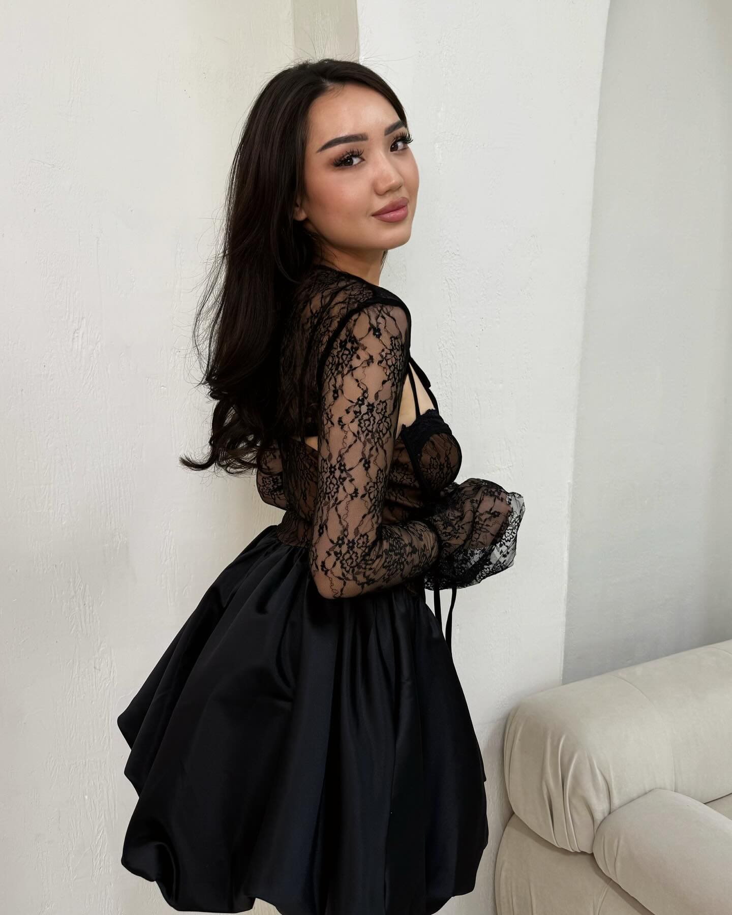 A woman stands in profile against a white wall, wearing a black lace long-sleeved top with a sheer back and a black satin skirt.