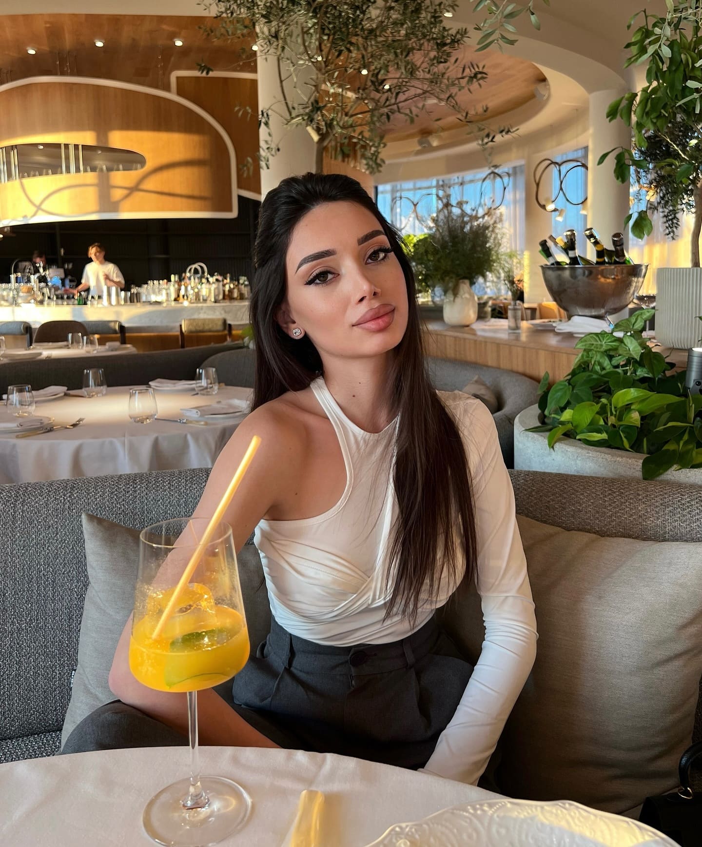 A woman in a white halter top and dark trousers sits at a restaurant table with a glass of orange drink in front of her.