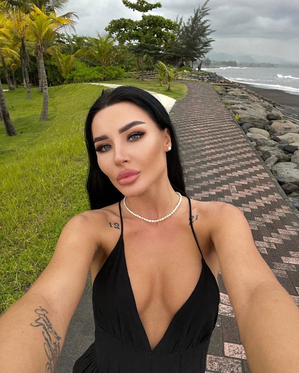 Victoria takes a coastal selfie in a black halter dress and pearl necklace, with palm trees and the ocean behind her.