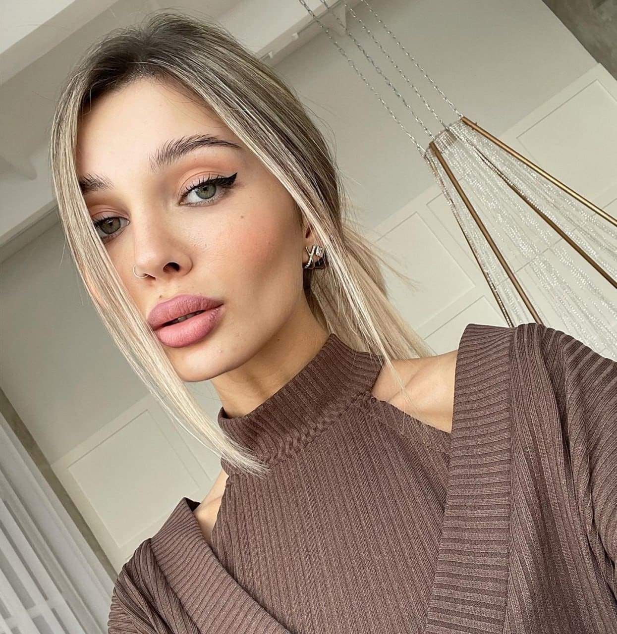Mila in a close-up selfie wearing a brown ribbed knit top with cut-out shoulders against a white paneled wall.