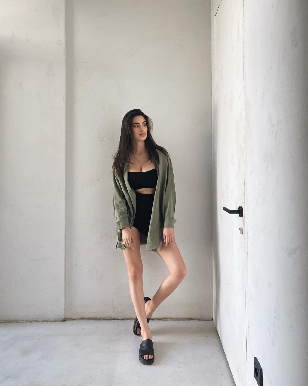 A woman stands in a white-walled corner wearing a black bandeau top, high-waisted shorts, and an unbuttoned olive-green shirt, with legs crossed.