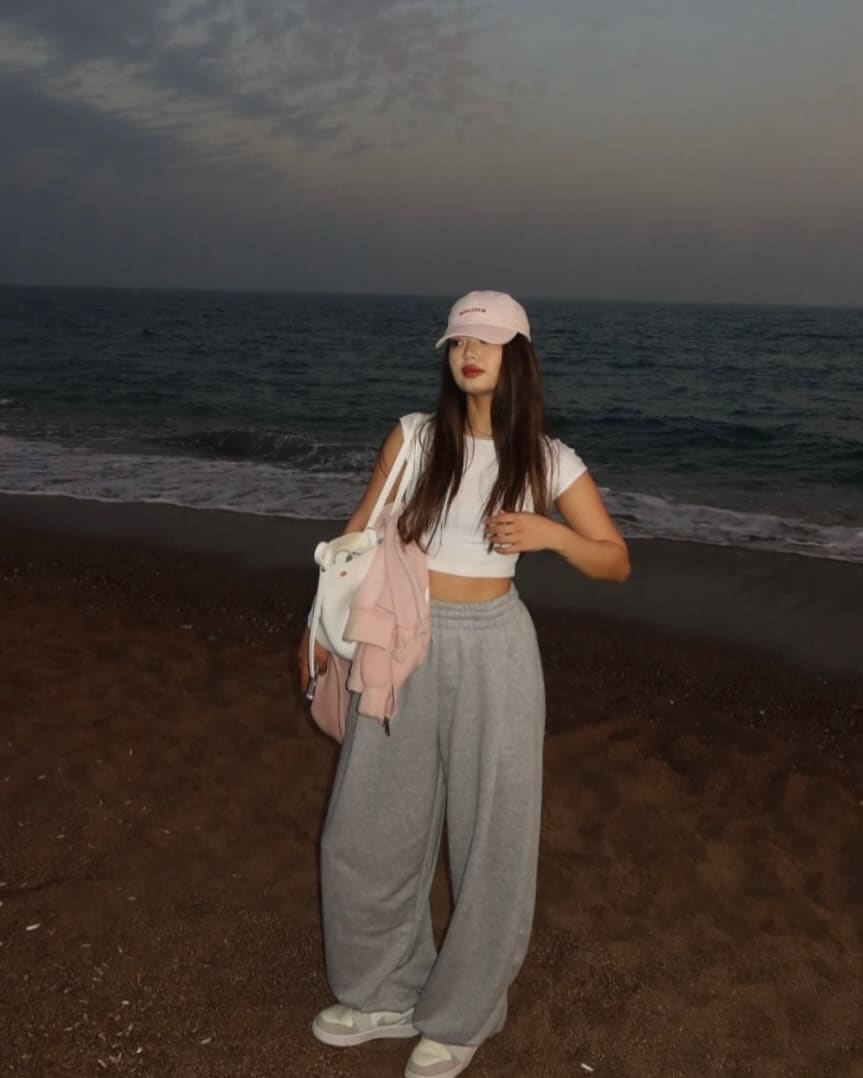 A woman in a pink cap, white crop top, and grey sweatpants stands on a dark-sand beach at dusk, ocean behind her.