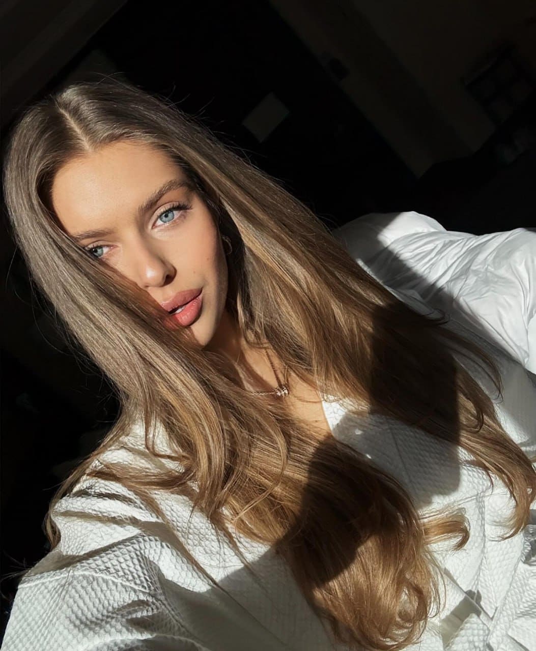 Nana lies on a white textured surface in a white top and thin necklace, sunlight casting shadows on her face against a dark background.