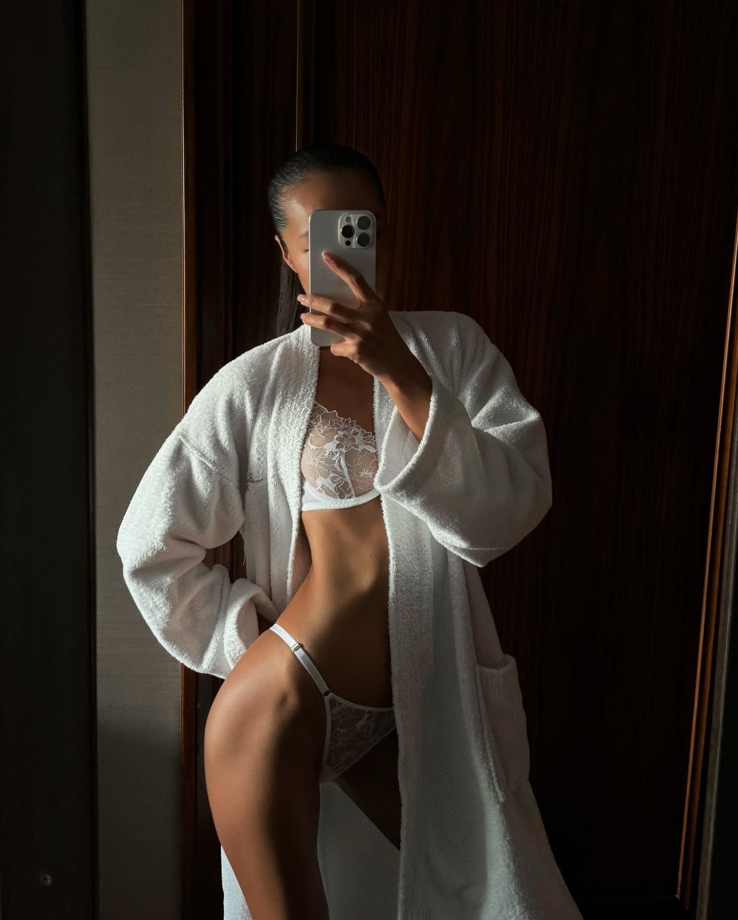 A mirror selfie shows a woman in an open white bathrobe and lace underwear against dark wood paneling, holding a phone to her face.