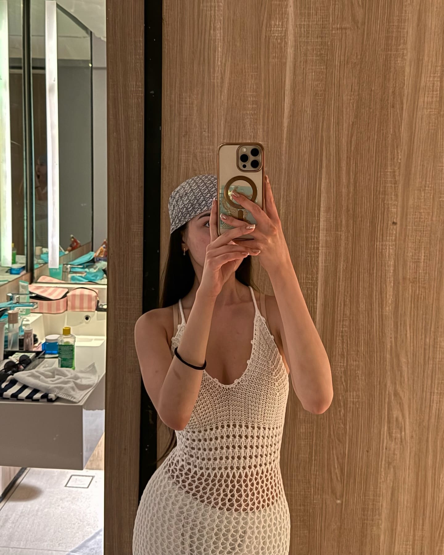 A mirror selfie shows a woman in a white crocheted halter dress and headscarf, holding a gold phone in a bathroom setting.
