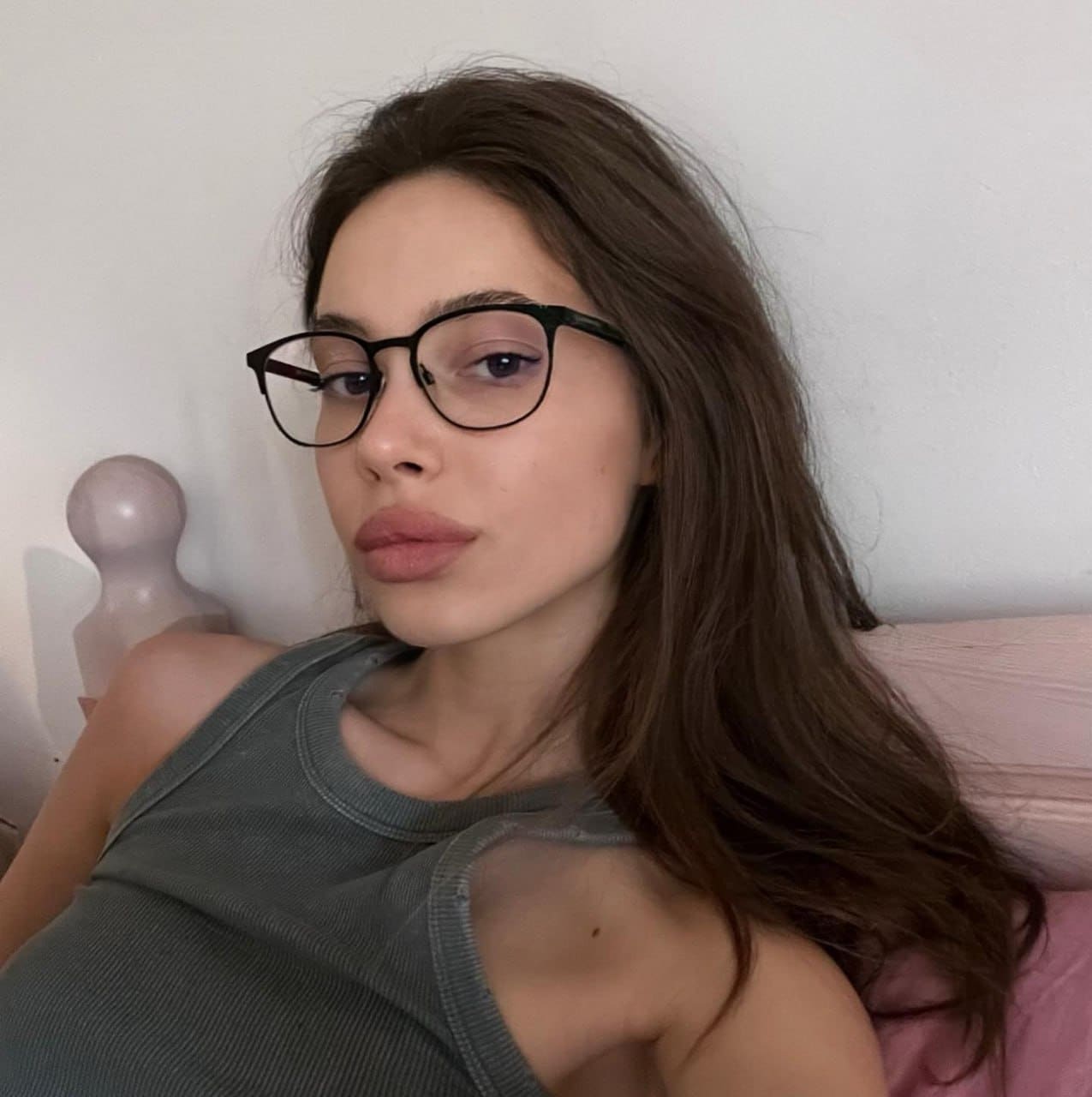 A woman with glasses lies on a bed with pink bedding, wearing a gray ribbed tank top with a shoulder cutout, looking at the camera.