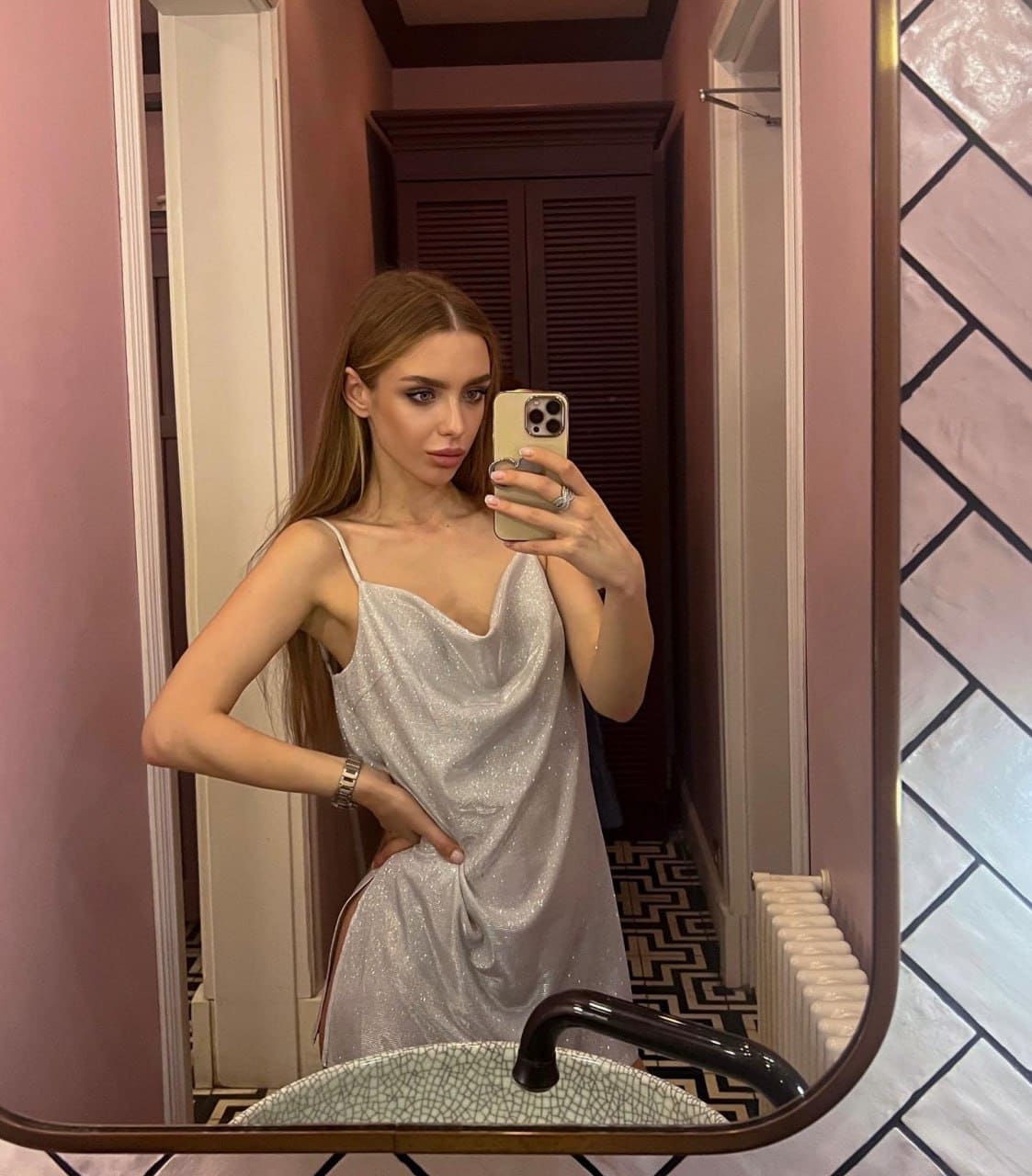 A mirror selfie shows a woman in a silver sparkly slip dress holding a phone, with a tiled sink in the background.