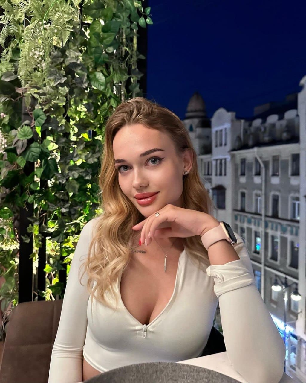 Anastasia sits at a table in a white zip-front top, resting her chin on her hand against a backdrop of green foliage and city buildings at dusk.