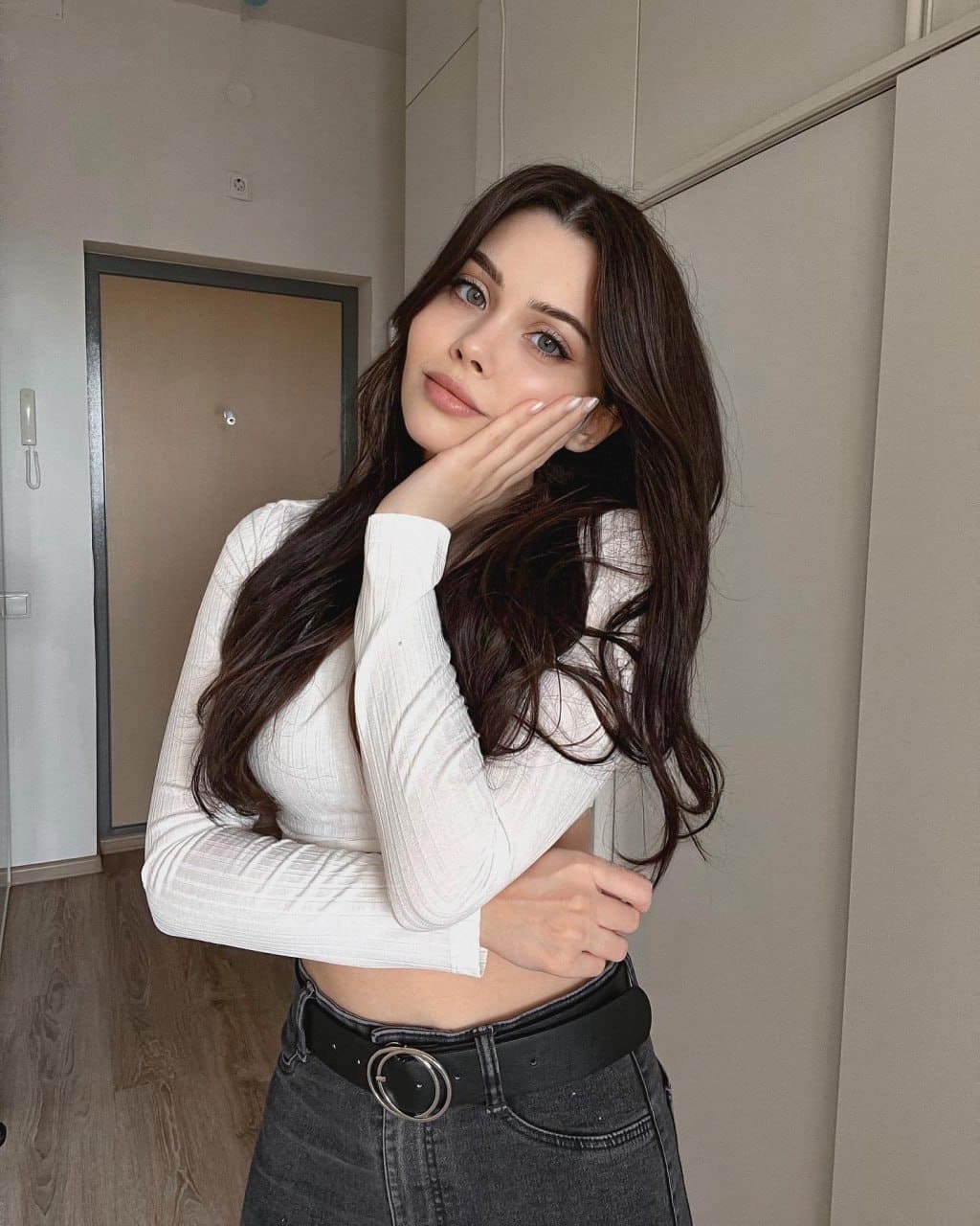 A woman in a white ribbed crop top and dark jeans stands indoors by a door, resting her right hand on her cheek.