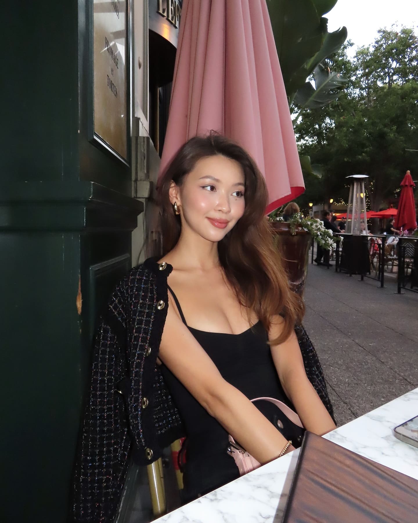 Gulnaz sits at an outdoor cafe table in a black spaghetti-strap top and tweed jacket, with a pink umbrella and foliage behind her.