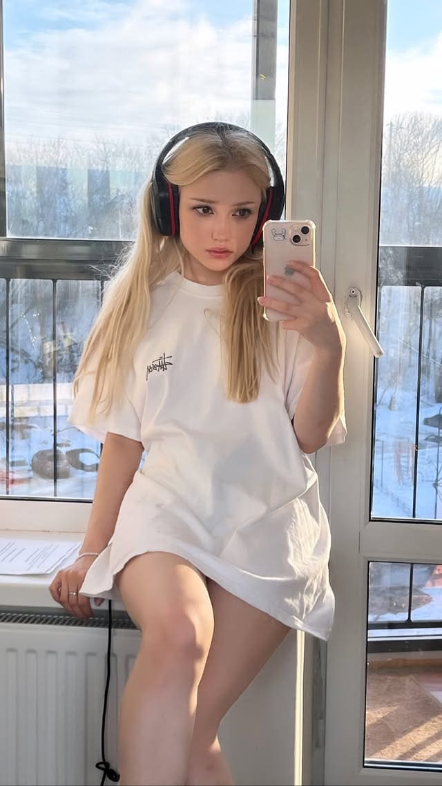 A blonde woman in a white t-shirt and headphones sits on a windowsill, holding a phone with snow visible outside.