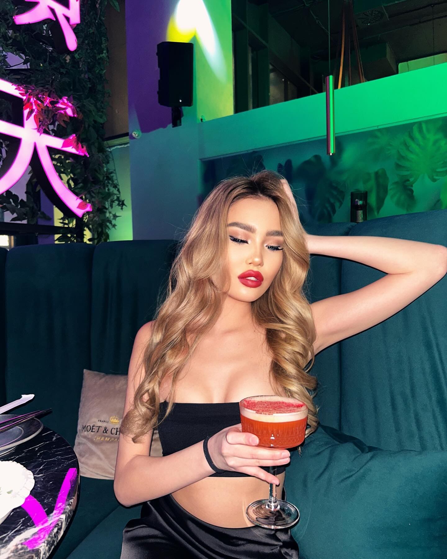 Florinel sits on a dark green couch in a dimly lit room with neon lighting, wearing a black strapless top and holding a stemmed glass with a red drink.