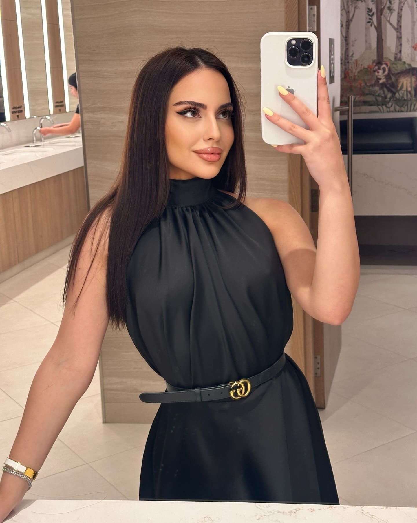 A mirror selfie in a bathroom shows a woman in a black high-neck dress with a thin belt, holding a white smartphone.