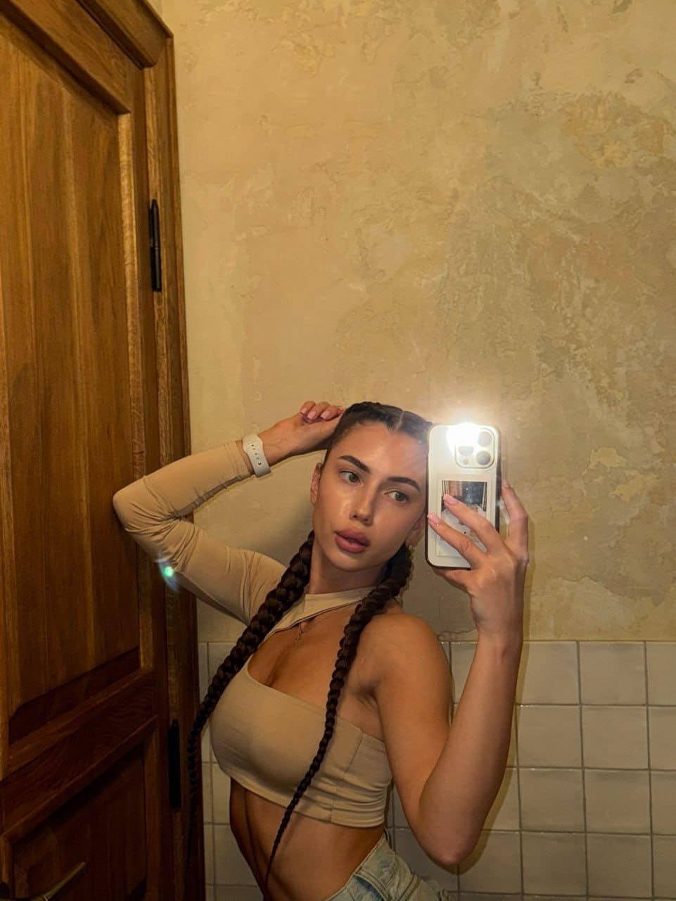 Bathroom mirror selfie in a beige crop top and denim shorts, one hand on head, against a textured wall and white tile wainscoting.