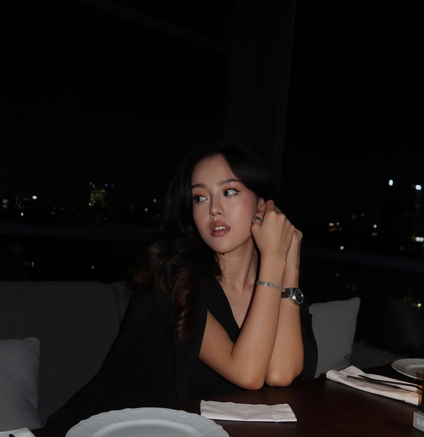 A woman in a black sleeveless top sits at a table in a dim room with hands clasped near her face, a city skyline visible through the window.