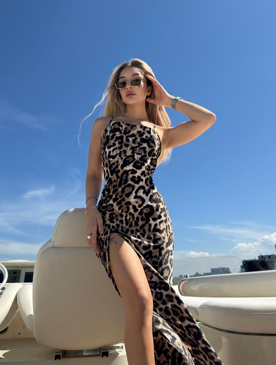 A blonde woman in a leopard-print dress and sunglasses stands on a boat, hand raised to her hair against a blue sky.